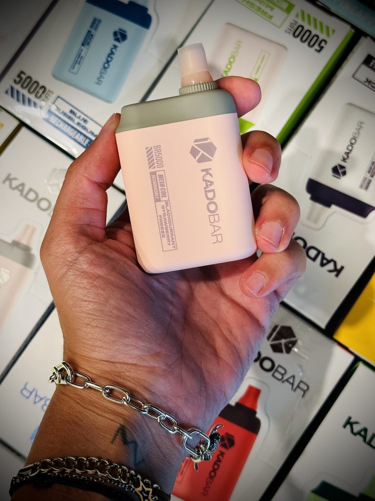 A close-up image of a person's hand holding a beige Kado Bar disposable vape device against a background of various Kado Bar packages. The hand is adorned with a silver chain bracelet and the vape has a minimalist design with the Kado Bar logo and product information on it. The surrounding boxes have "5000 PUFFS" prominently displayed, indicating the product's usage capacity.