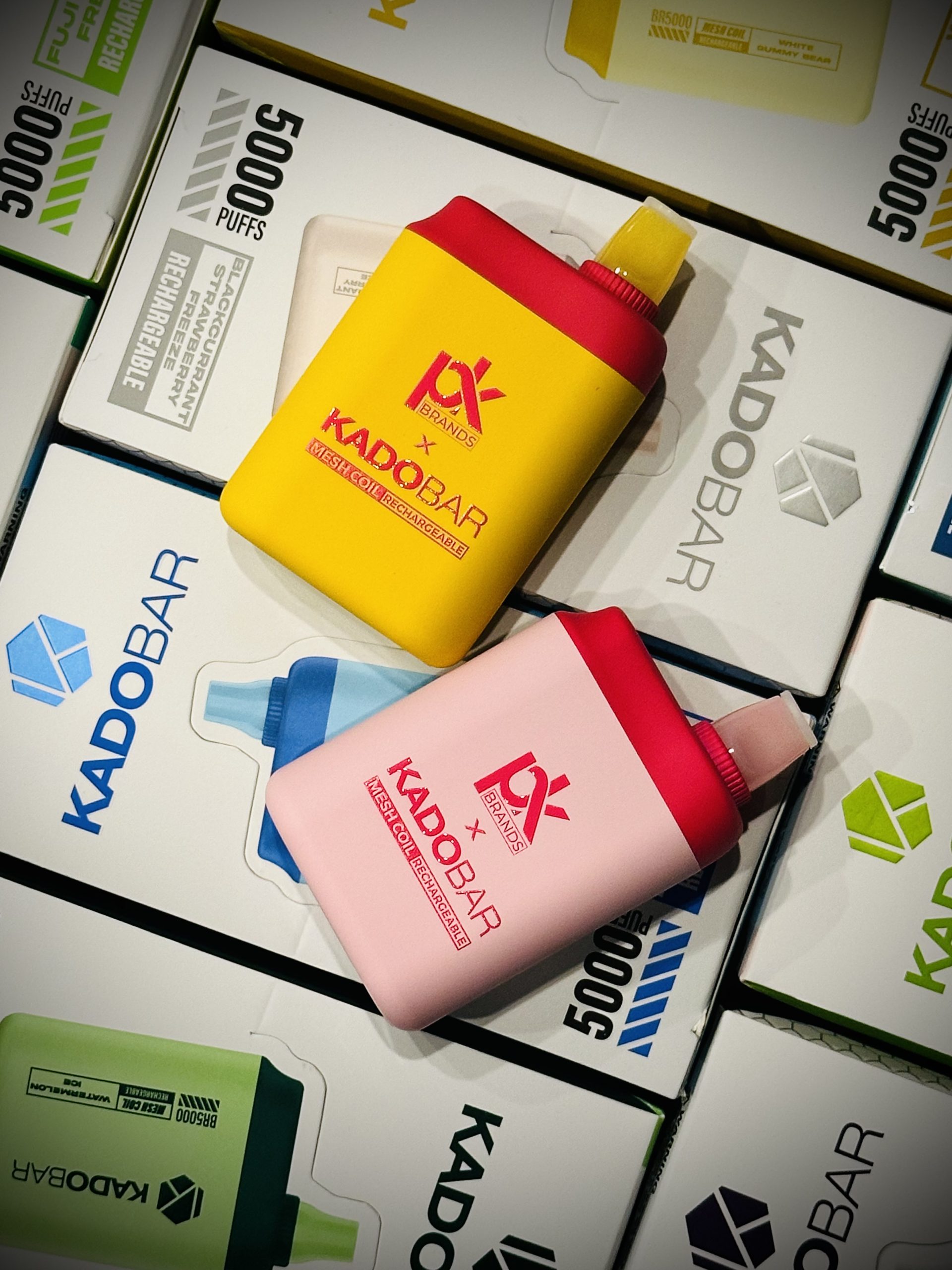An image of two Kado Bar disposable vape devices in yellow and pink, prominently displayed among various packaging boxes. The boxes have a clean design with the text 